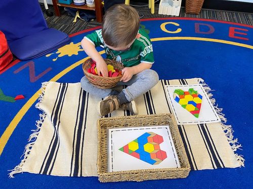 A student sitting on the floor sorts through assorted tiles to match them to an print out that outlines those shapes to match.