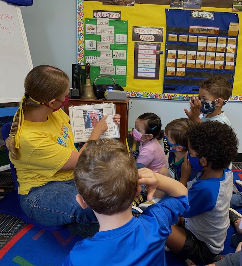 Ms. Mieke sitting on the ground holding with children gathered around her looking at the book she is holding in the classroom.